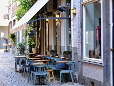 A sunny terrace surrounded by buildings and cafes. Blue chairs and wooden tables are arranged in front of the café.