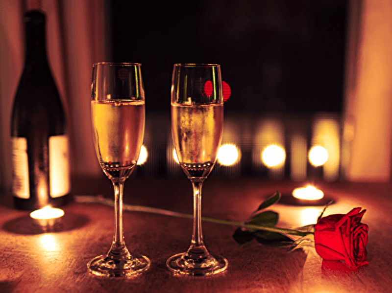 Twee gevulde champagneglazen op een houten tafel met een rode roos en brandende kaarsjes, op de achtergrond een fles wijn en zachte sfeerverlichting voor een romantische setting.