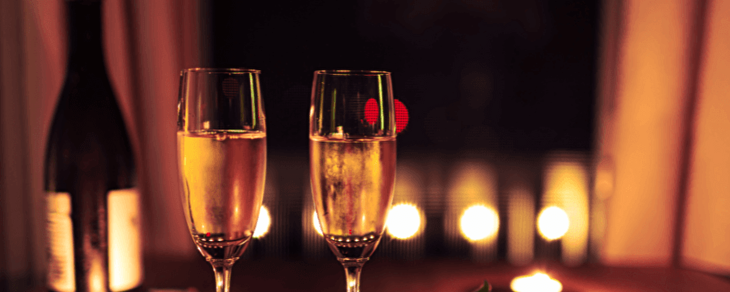 Twee gevulde champagneglazen op een houten tafel met een rode roos en brandende kaarsjes, op de achtergrond een fles wijn en zachte sfeerverlichting voor een romantische setting.