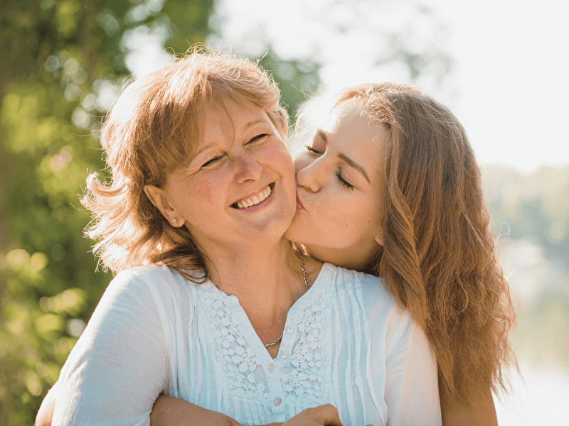 A smiling mother is lovingly embraced and kissed on the cheek by her daughter during a sunny day in a green, natural setting.